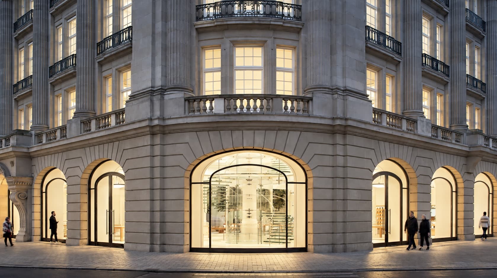 Leidseplein'deki Apple Amsterdam, alacakaranlıkta; aydınlatılmış kemerli pencereler ve cam bir merdiven